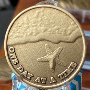 Pode incluir: Uma moeda redonda, cor de bronze, com uma cena de praia. A moeda apresenta uma estrela-do-mar na areia e a frase "ONE DAY AT A TIME". A moeda tem uma superf&iacute;cie texturizada.