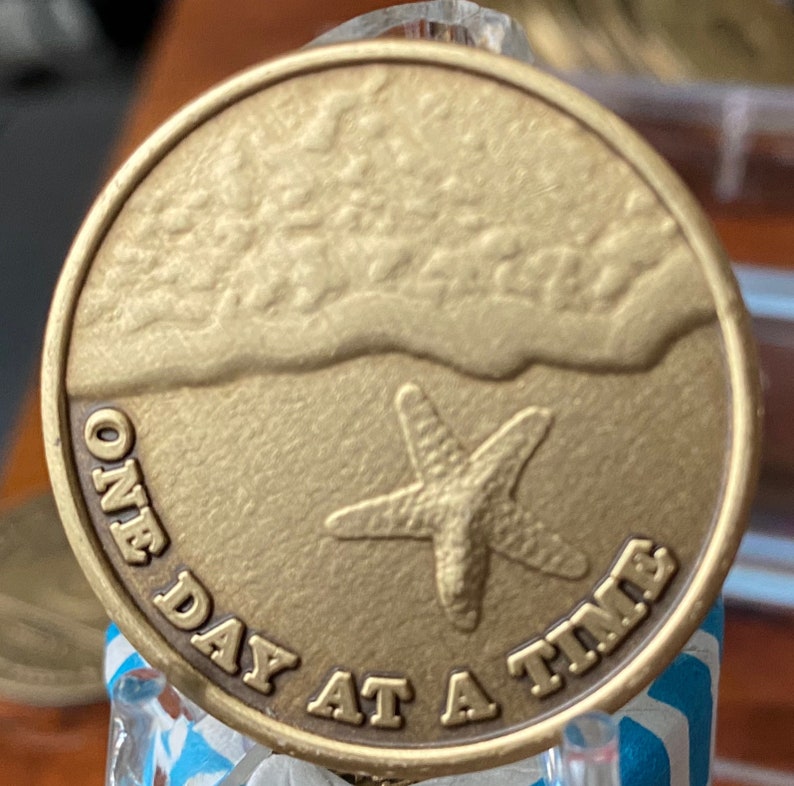 Pode incluir: Uma moeda redonda, cor de bronze, com uma cena de praia. A moeda apresenta uma estrela-do-mar na areia e a frase "ONE DAY AT A TIME". A moeda tem uma superf&iacute;cie texturizada.