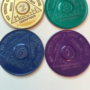 May include: Four gold, green, blue, and purple metal sobriety coins with a triangle design. Each coin has a number, "Month", and "Recovery" inscribed on it. The coins are for 2, 3, 6, and 9 months of sobriety.