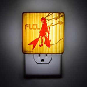 Puede incluir: Una luz nocturna amarilla con una silueta roja de una persona sosteniendo una guitarra. La luz nocturna tiene el texto "FLCL" y el texto "boyDesigns" en la parte inferior.