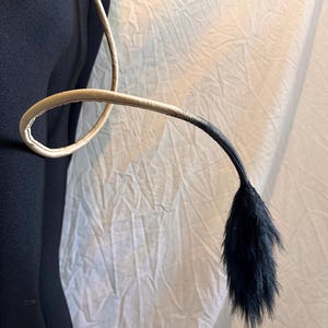 May include: A costume tail featuring a cream-coloured, stitched leather-like loop and a black, fluffy tail. The tail transitions from cream to black. The tail is likely for cosplay or costume use.