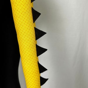 Pu&ograve; includere: Un tubo di tessuto giallo con un motivo a rombi e punte triangolari nere lungo un bordo. Il tubo &egrave; posto su uno sfondo nero e uno sfondo bianco.
