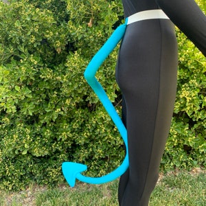 Peut inclure: Une queue de diable bleu clair, incurvée et pointue à l'extrémité, attachée à une tenue noire avec une ceinture blanche. La queue est d'un bleu vif, contrastant avec les vêtements sombres. L'arrière-plan est un buisson vert.