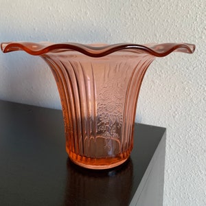 Pode incluir: Um vaso de vidro rosa com uma borda festonada e um design floral. O vaso tem uma textura canelada e mede aproximadamente 15 cm de altura.