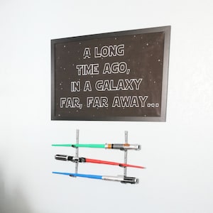 May include: A blue bedroom with a bed, a white headboard, and a navy blue comforter. There are three framed prints on the wall behind the bed, and a black framed chalkboard with the text "A long time ago, in a galaxy far, far away..." on the wall to the right of the bed. There are three lightsabers hanging on the wall below the chalkboard.