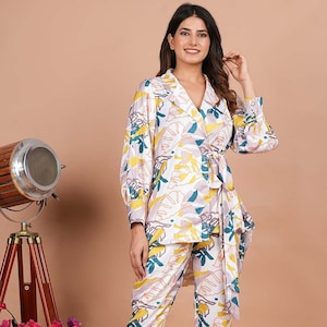 Op de afbeelding: Een wit, roze en geel geprint broekpak met een lang mouw shirt en bijpassende broek. Het shirt heeft een strik in de taille en een kraag.