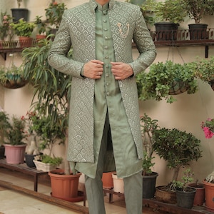 May include: A sage green long-sleeved jacket with a geometric pattern, worn over a matching sage green shirt and pants. The jacket has a mandarin collar and a button closure. The man is wearing brown leather loafers.