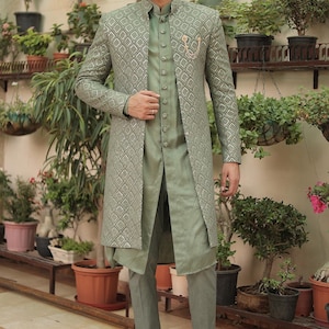 May include: A green patterned long jacket with a matching green shirt and pants. The jacket has a silver thread design and a gold button closure. The man is wearing brown loafers.