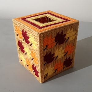 May include: A handmade tissue box cover featuring a fall leaf design. The box is square, with a tan base color and a pattern of orange, yellow, and burgundy leaves. The top has a square opening with a decorative border.
