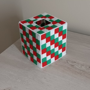 May include: A square tissue box cover with a checkerboard pattern in red, green, and white. The box has a square opening on top for tissue dispensing. The cover is made of a woven material and sits on a light-colored surface.