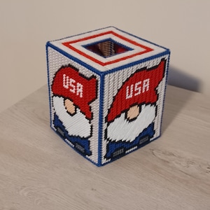 May include: A square tissue box cover featuring a pixelated design of a gnome wearing a red hat with "USA" on it. The box has a white background with blue and red trim. The gnome has a white beard and blue pants.