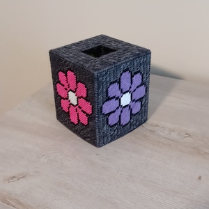 May include: A square tissue box cover made of dark gray plastic canvas. The cover features a pink and a purple flower design with white centers. The tissue box cover is sitting on a light wood surface.