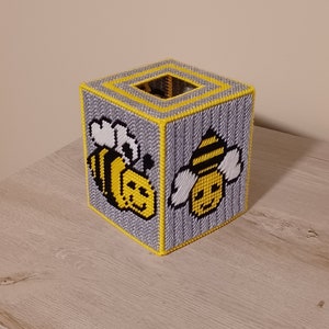 May include: A handmade tissue box cover in a cube shape. The box is gray with yellow trim and features two smiling bee designs. The bees are yellow and black with white wings. The tissue box cover is made of plastic canvas.
