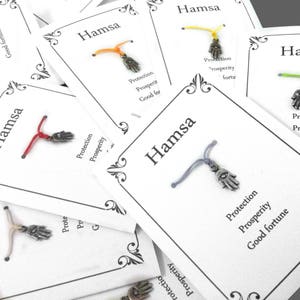 May include: A set of six Hamsa hand charm bracelets with different colored strings. Each bracelet has a silver charm with the Hamsa hand symbol and the words "Protection, Prosperity, Good fortune" printed on the card.
