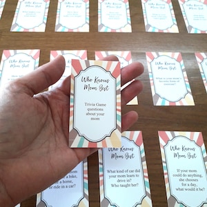 May include: A set of colorful trivia game cards with the title "Who Knows Mom Best?" and questions about a person's mother. The cards have a striped background with a white border and a decorative frame.