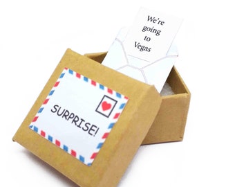 Peut inclure: Une petite boîte en carton marron avec un cœur rouge et les mots "SURPRISE!" sur le devant. La boîte est ouverte et une enveloppe blanche avec les mots "We're going to Vegas" est à l'intérieur.