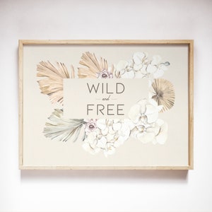 May include: A framed print featuring a watercolor floral arrangement with the words "WILD and FREE". The artwork includes beige, white, and light brown flowers and leaves, set against a neutral background. The frame is made of light-colored wood.
