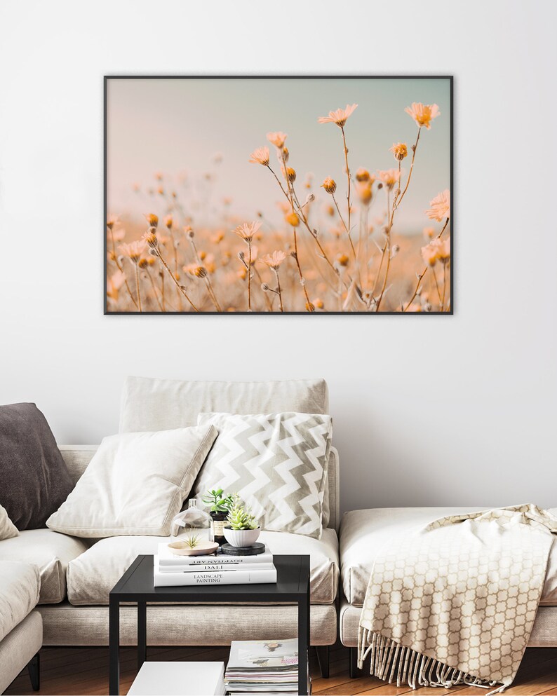 Pu&ograve; includere: Una stampa incorniciata con un campo di delicati fiori selvatici color pesca contro un cielo azzurro tenue. L'opera d'arte &egrave; esposta sopra un divano beige con cuscini e un tavolino nero.