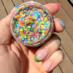 May include: A clear, round container filled with colorful glitter. The glitter pieces are various shapes and sizes, including hexagons, and in shades of pink, yellow, blue, and green. The container is held in a hand with painted nails.