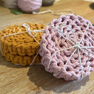 May include: Two stacks of handmade crochet coasters. One stack is mustard yellow, the other is light pink. Each stack is tied with white twine. The coasters are on a wooden surface.