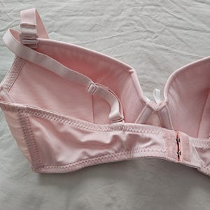 May include: A light pink bra with adjustable straps and a hook-and-eye closure. The bra features a classic design with seamed cups and a soft, smooth fabric. The straps and closure are also light pink.