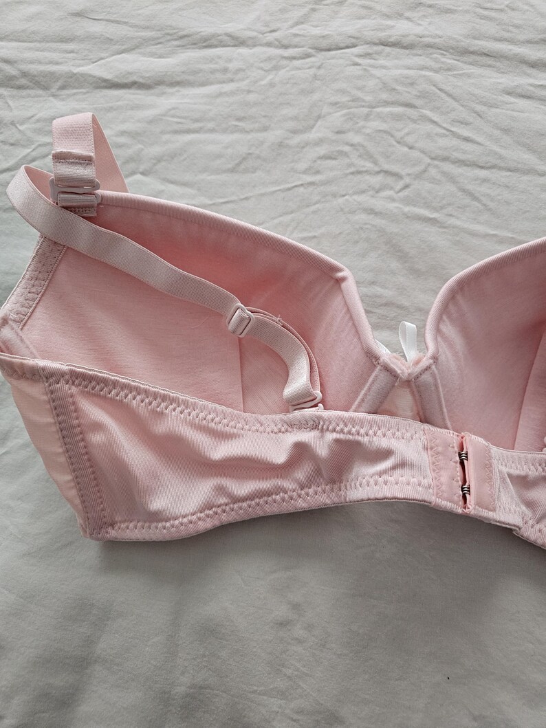 May include: A light pink bra with adjustable straps and a hook-and-eye closure. The bra features a classic design with seamed cups and a soft, smooth fabric. The straps and closure are also light pink.