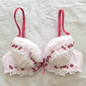 May include: A light pink underwire bra featuring white lace overlays on the cups, accented with small dark pink floral embroidery and ruffled trim. The garment includes adjustable dark pink shoulder straps and a small dark pink bow at the center.