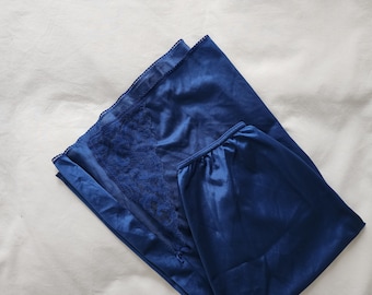May include: A navy blue satin pajama set. The top features a lace panel and decorative details. The set is made of a shiny, smooth fabric. The pajama set is folded on a white surface.
