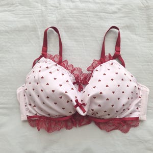 May include: A white bra with a pattern of small red hearts. The bra has red straps, trim, and lace detailing. A small red bow is centered between the cups. The bra is made of a soft, silky material.