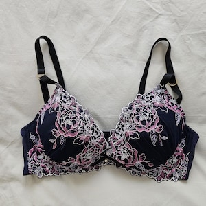 Könnte beinhalten: Ein schwarzer Spitzen-BH mit rosa Blumen Stickerei. Der BH hat verstellbare Träger und einen Haken- und Ösenverschluss auf der Rückseite.
