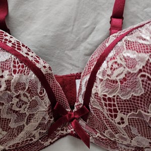 May include: A burgundy bra with white lace overlay. The bra features adjustable straps and a small bow detail at the center. The lace design is intricate, and the bra appears to be made of soft materials.