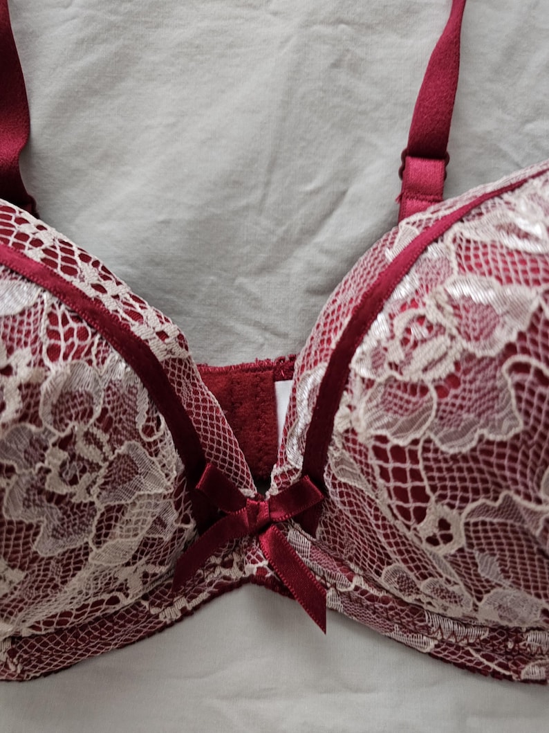 May include: A burgundy bra with white lace overlay. The bra features adjustable straps and a small bow detail at the center. The lace design is intricate, and the bra appears to be made of soft materials.