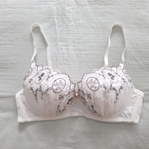 Op de afbeelding: Witte beha met delicate bloemenborduursels in zwart en roze. De beha heeft verstelbare bandjes en een geschulpte rand. Een klein roze strikje is gecentreerd tussen de cups.