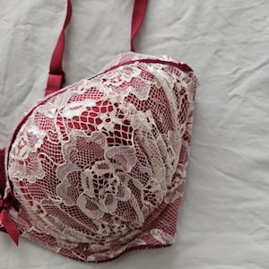 May include: A red bra with white lace overlay. The bra has adjustable straps and a clasp closure. A tag with the text "E70" is attached to the strap. The bra is made of a soft, delicate fabric.
