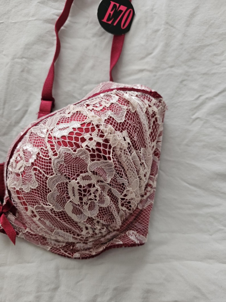 May include: A red bra with white lace overlay. The bra has adjustable straps and a clasp closure. A tag with the text "E70" is attached to the strap. The bra is made of a soft, delicate fabric.
