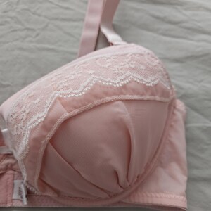May include: A light pink bra with white lace detailing along the top edge of the cups. The bra has adjustable straps and a hook-and-eye closure. The fabric appears to be a soft, smooth material.