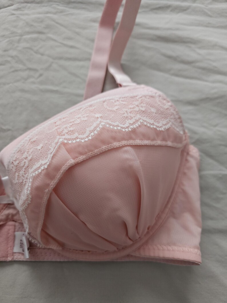 May include: A light pink bra with white lace detailing along the top edge of the cups. The bra has adjustable straps and a hook-and-eye closure. The fabric appears to be a soft, smooth material.