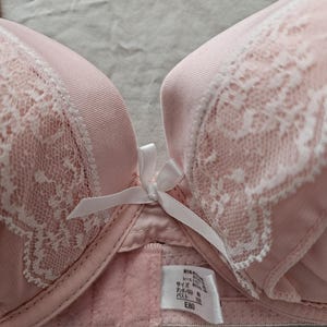 May include: A light pink bra with white lace detailing and a small white bow. The bra has a smooth, satin-like texture on the cups and straps. A tag with Japanese text is visible.