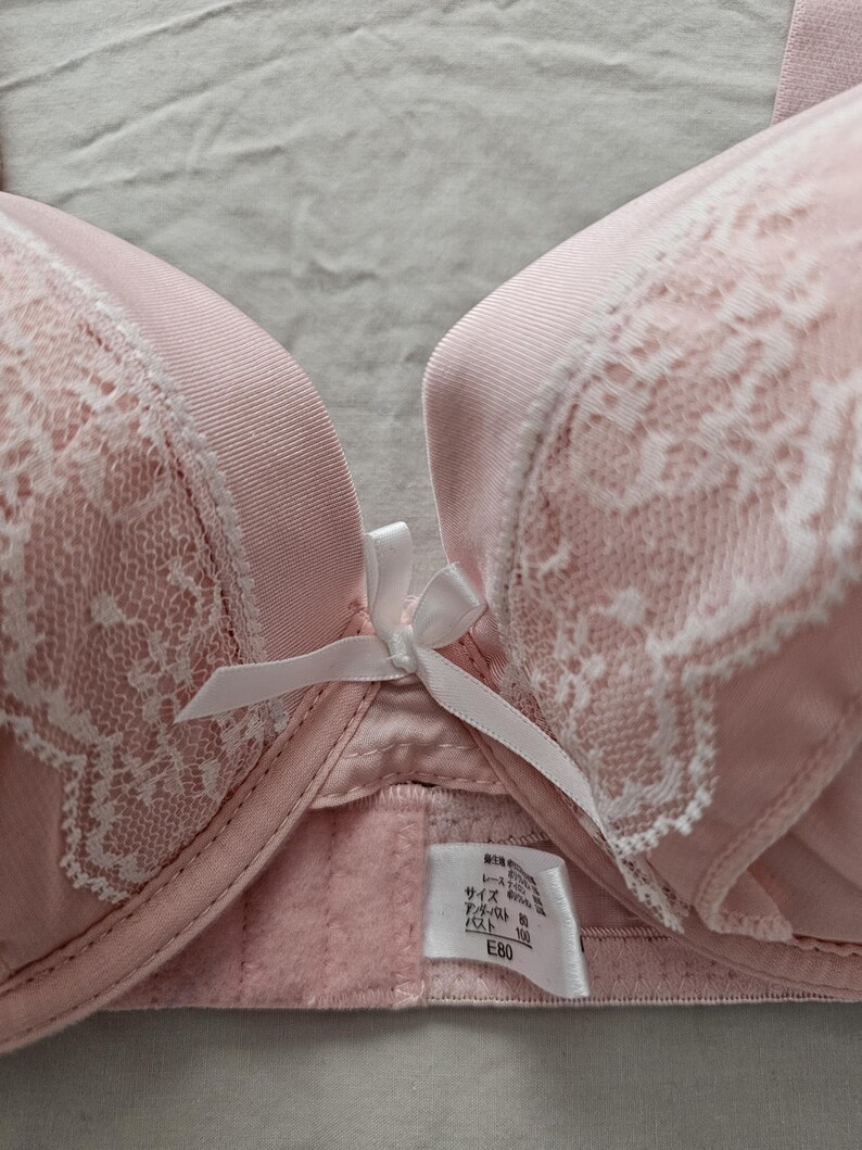 May include: A light pink bra with white lace detailing and a small white bow. The bra has a smooth, satin-like texture on the cups and straps. A tag with Japanese text is visible.