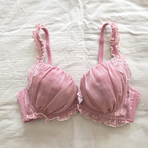 Puede incluir: Un par de sujetadores rosas con rayas verticales y detalles de encaje blanco. El sujetador presenta bordes con volantes y tirantes ajustables. El diseño es delicado y femenino, hecho de un material suave y sedoso.