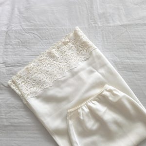 Op de afbeelding: Ivoor kleurige pyjama set. De top heeft een delicate kanten rand langs de halslijn. De set is gemaakt van een glad, zijdeachtig materiaal en is netjes op een wit oppervlak gevouwen. De algehele esthetiek is zacht en elegant.