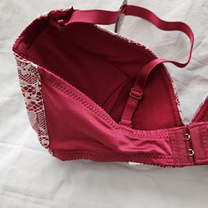 May include: A burgundy bra with white lace detailing along the edges. The bra features adjustable straps and a hook-and-eye closure. The bra is made of a smooth, satin-like material.