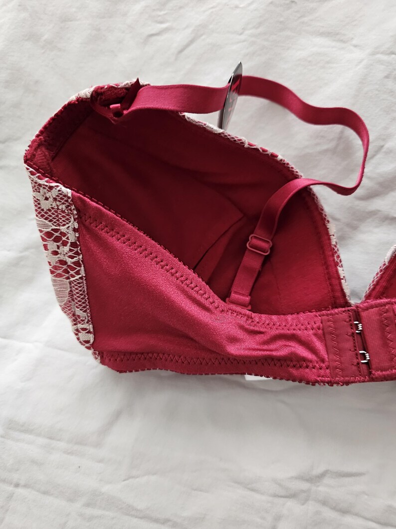 May include: A burgundy bra with white lace detailing along the edges. The bra features adjustable straps and a hook-and-eye closure. The bra is made of a smooth, satin-like material.
