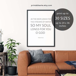 May include: A printable wall art design featuring a black framed poster with a white background and the text "As the deer longs for the streams of water, so my soul longs for you O God. Psalm 42:1". The poster is displayed on a brown leather couch with a gray throw blanket and a small table with a book on it. The text "print up to 30 sizes up to 24 x 36 inches" is displayed in a white circle with a black outline.