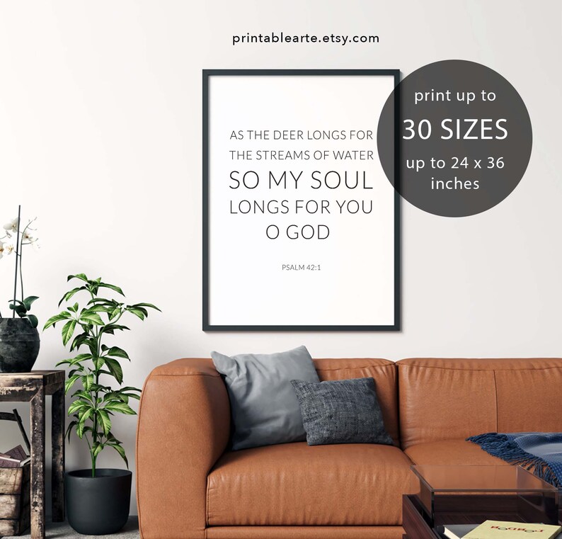 May include: A printable wall art design featuring a black framed poster with a white background and the text "As the deer longs for the streams of water, so my soul longs for you O God. Psalm 42:1". The poster is displayed on a brown leather couch with a gray throw blanket and a small table with a book on it. The text "print up to 30 sizes up to 24 x 36 inches" is displayed in a white circle with a black outline.