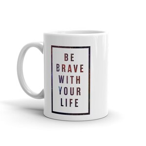 Pode incluir: Caneca de caf&eacute; de cer&acirc;mica branca com uma borda preta em torno de uma impress&atilde;o de gal&aacute;xia. O texto "BE BRAVE WITH YOUR LIFE" &eacute; impresso em preto na caneca.