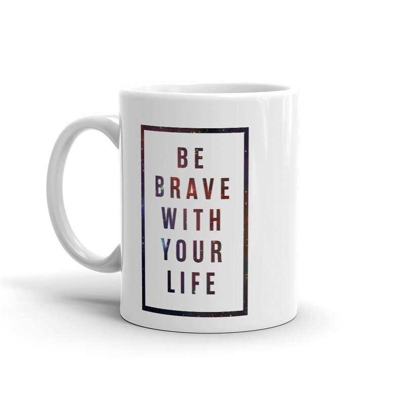 Pode incluir: Caneca de caf&eacute; de cer&acirc;mica branca com uma borda preta em torno de uma impress&atilde;o de gal&aacute;xia. O texto "BE BRAVE WITH YOUR LIFE" &eacute; impresso em preto na caneca.