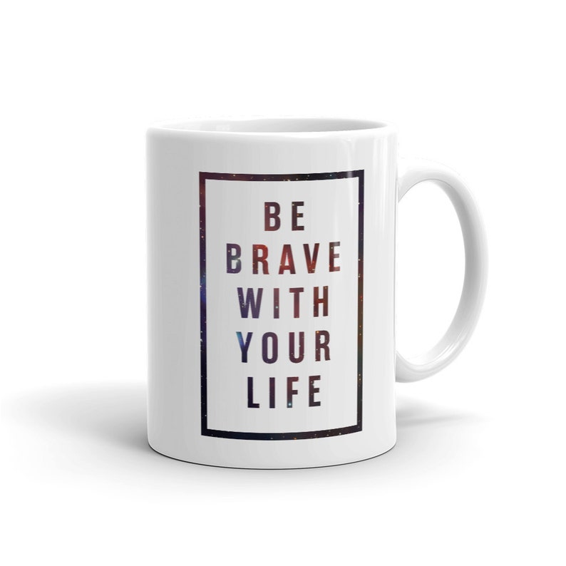 Pode incluir: Caneca de cer&acirc;mica branca com uma borda preta em torno do texto "BE BRAVE WITH YOUR LIFE" impresso em preto sobre um fundo de gal&aacute;xia.
