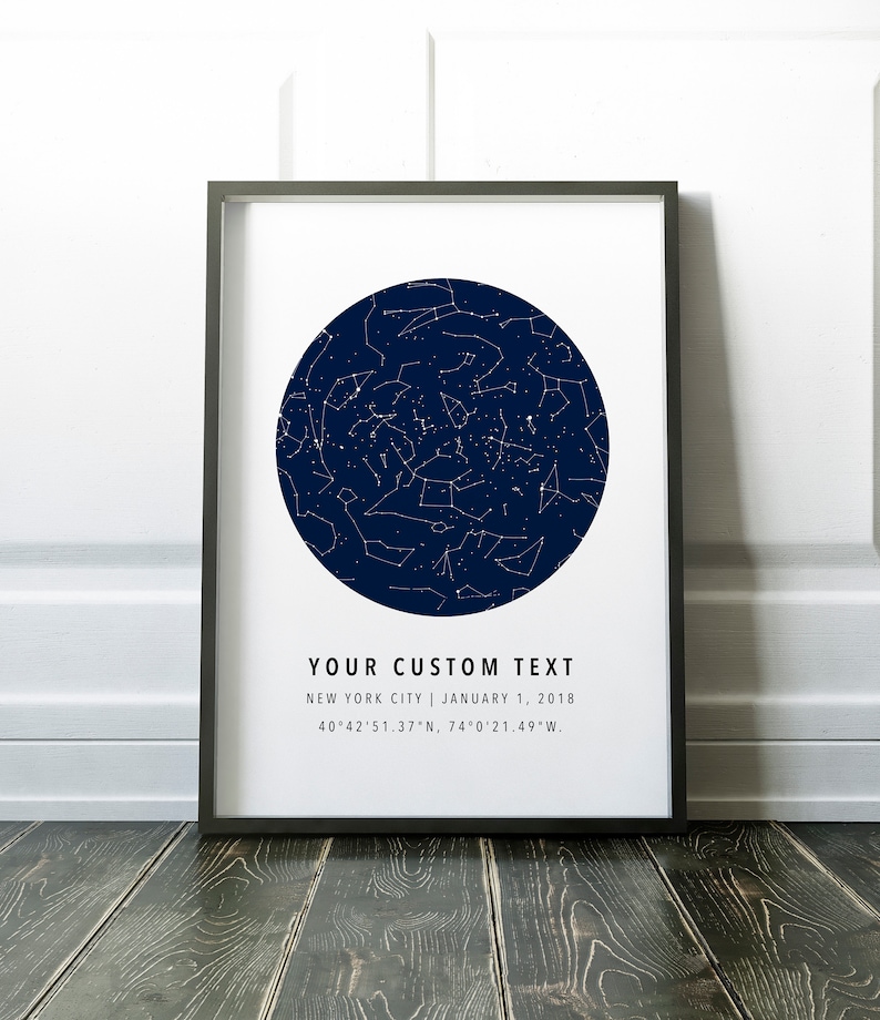 May include: Framed print featuring a dark blue circular star map with white constellations. The print includes the text "YOUR CUSTOM TEXT", location, and date. The frame is black, and the print is displayed on a wooden surface.