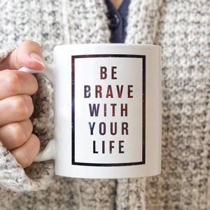 Pode incluir: Caneca de cer&acirc;mica branca com uma borda preta em torno de um fundo semelhante a uma gal&aacute;xia. O texto "BE BRAVE WITH YOUR LIFE" &eacute; impresso em preto.